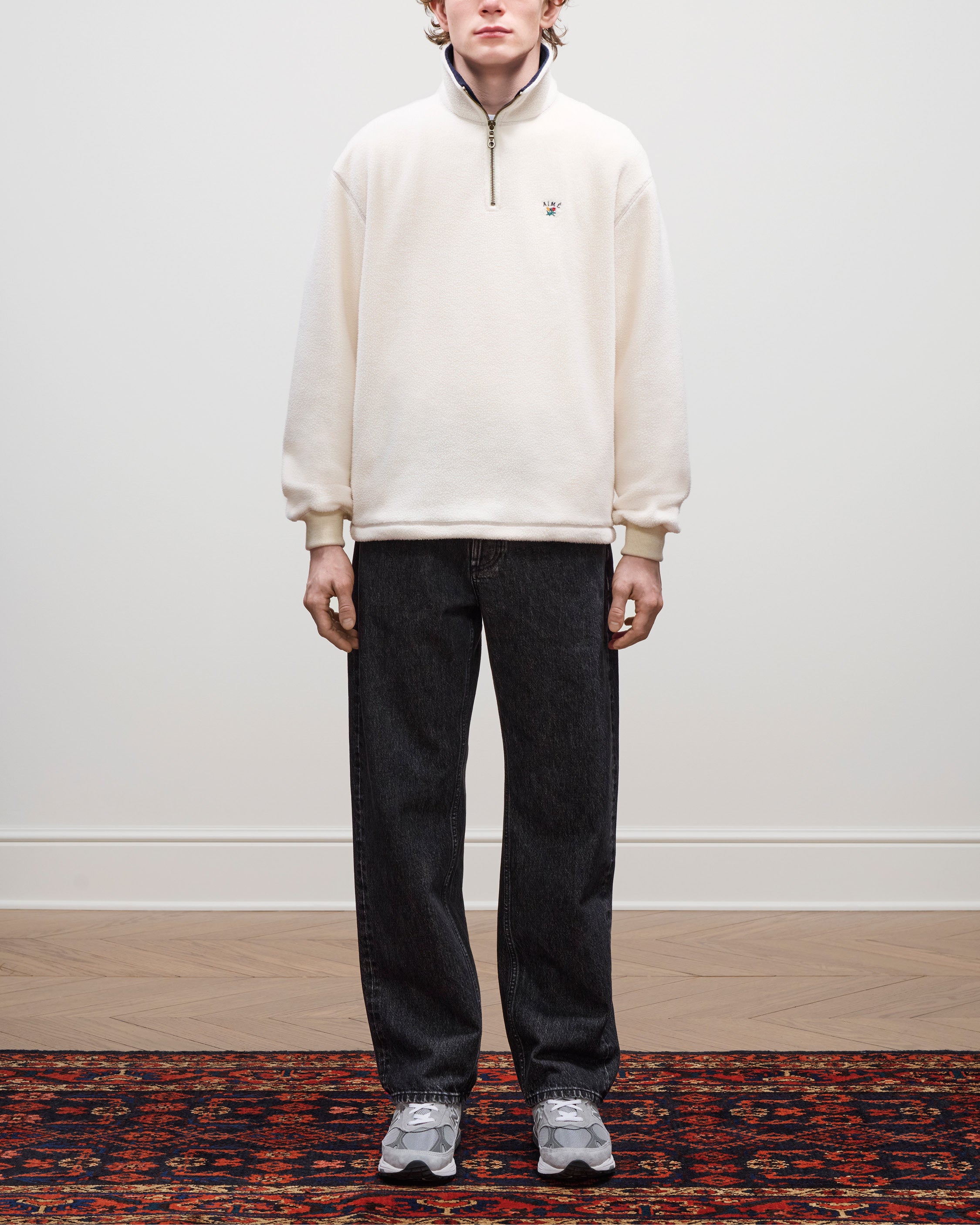 A person stands indoors wearing a Crest Quarter-Zip Fleece Pullover, dark jeans, and gray sneakers on a patterned rug.