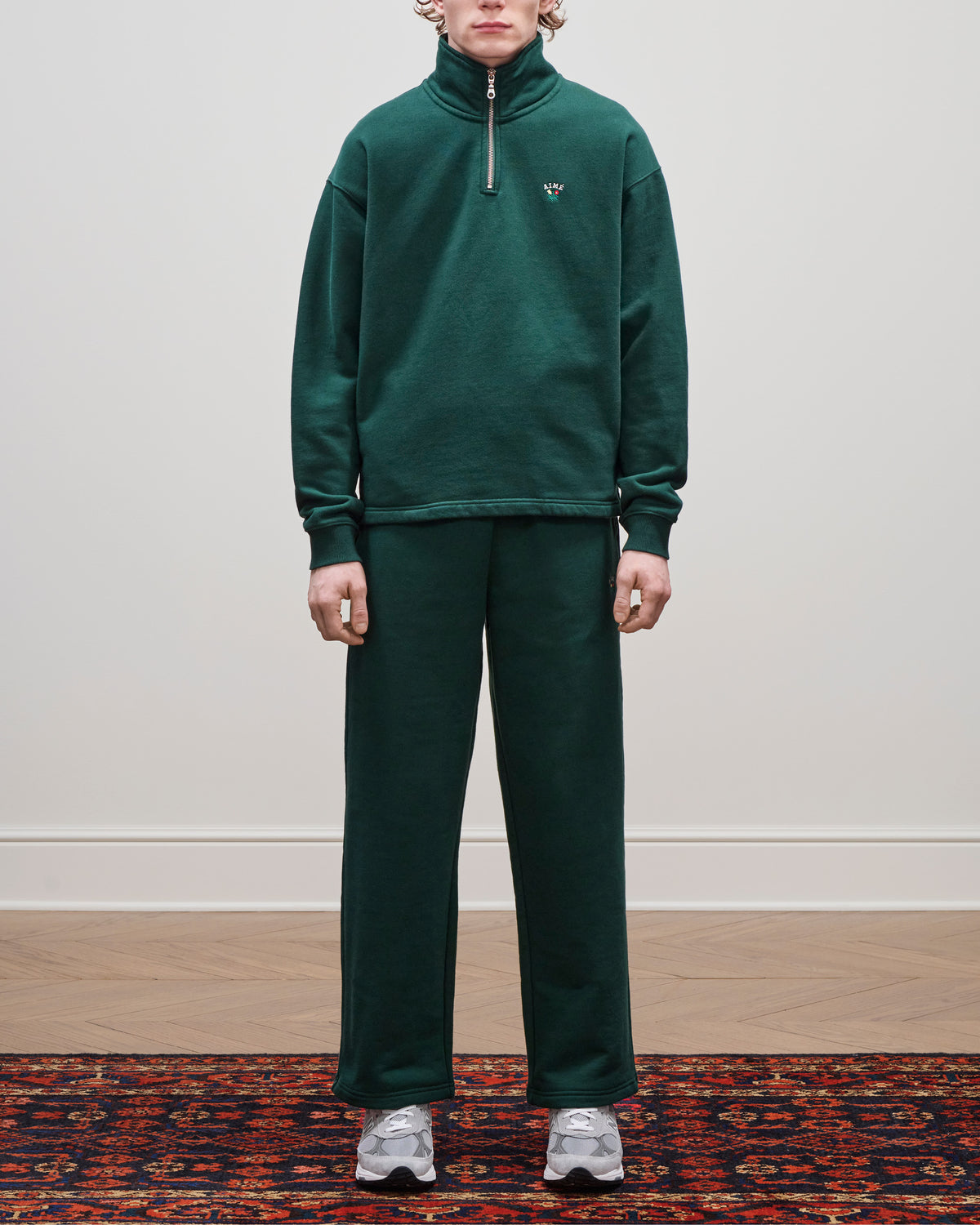 Person in a Crest Quarter Zip Pullover and gray sneakers stands on a patterned rug against a plain wall.