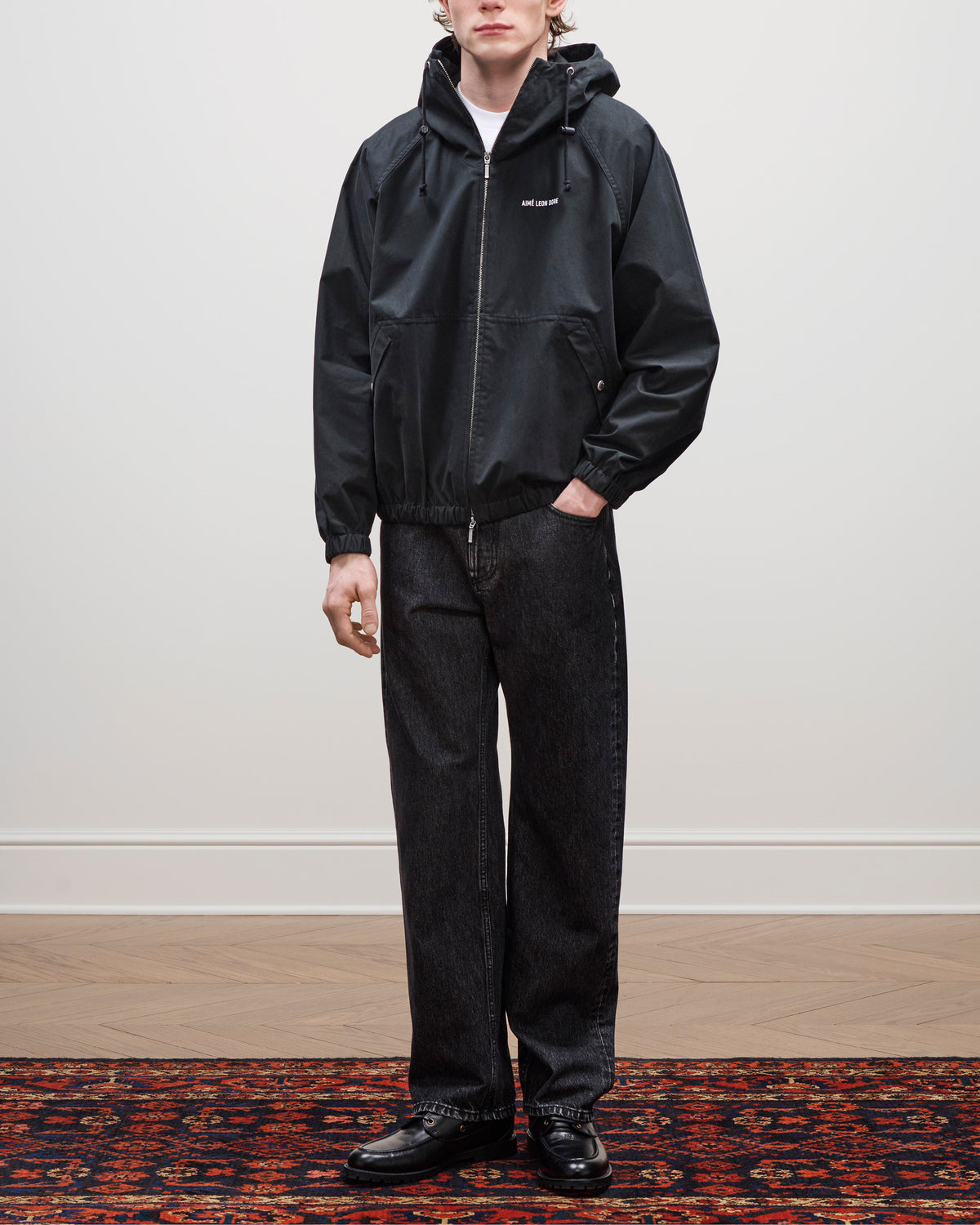Person wears the Micro Logo Lightweight Jacket, dark jeans, and black shoes on a patterned rug against a plain wall.
