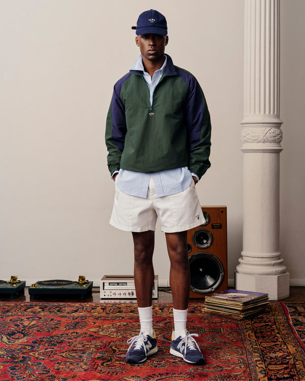 A man in a Crest Nylon Half-Zip Pullover, light blue shirt, white shorts, and sneakers stands on a rug indoors.