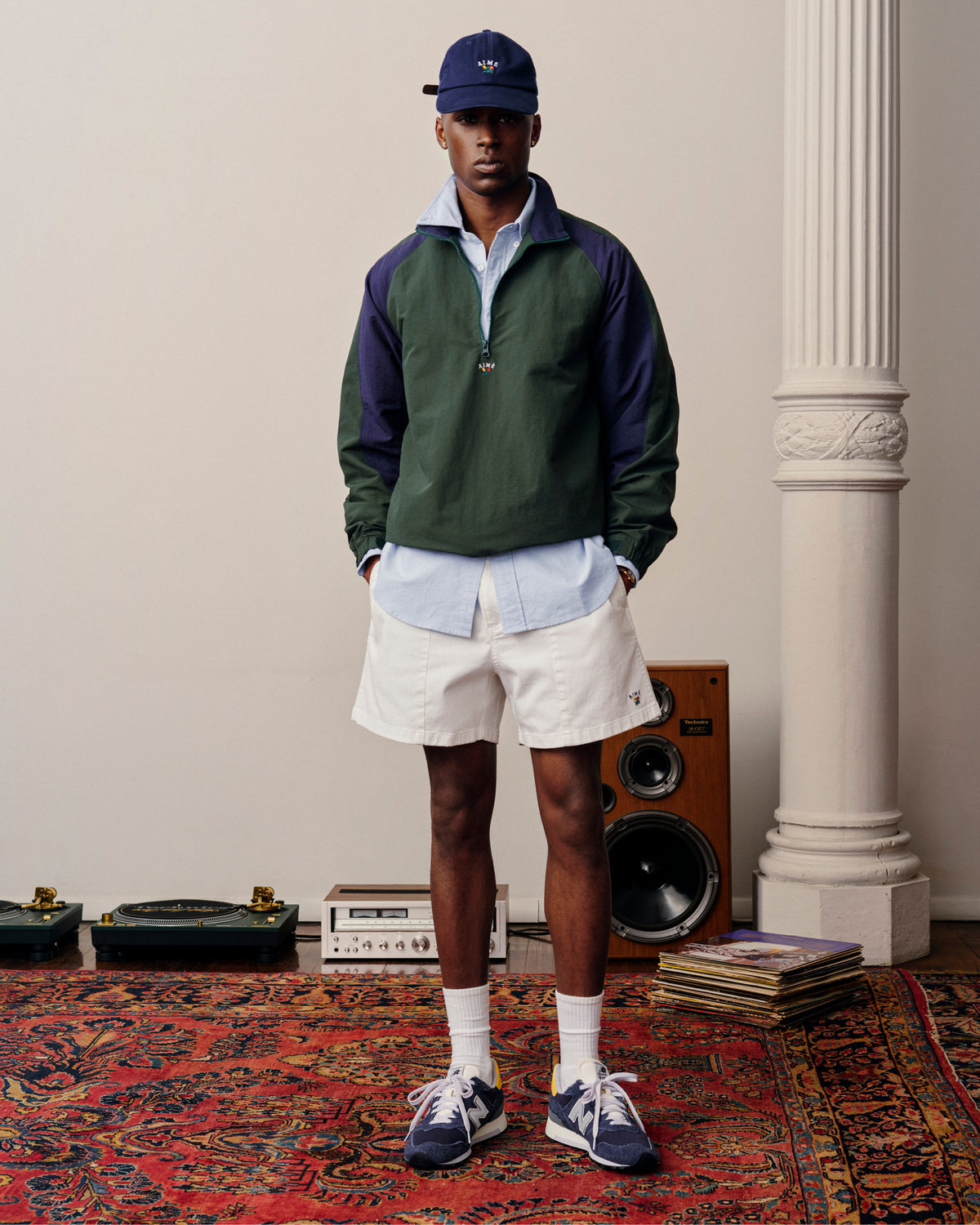 A man in Crest Casual Shorts, a green and navy jacket, and blue cap stands on a red patterned rug near speakers and records.