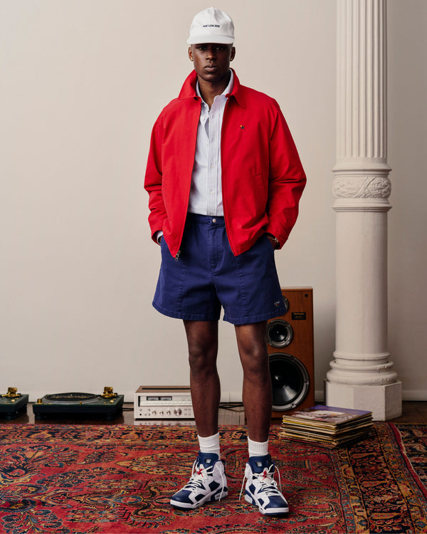 A man in a Crest Casual Jacket, navy shorts, and white cap stands on a patterned rug beside speakers and vinyl records.