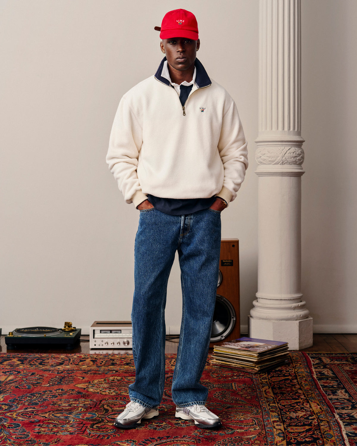 A man in a white pullover, blue jeans, Crest Logo Hat, and silver sneakers stands on a patterned rug in a stylish room.
