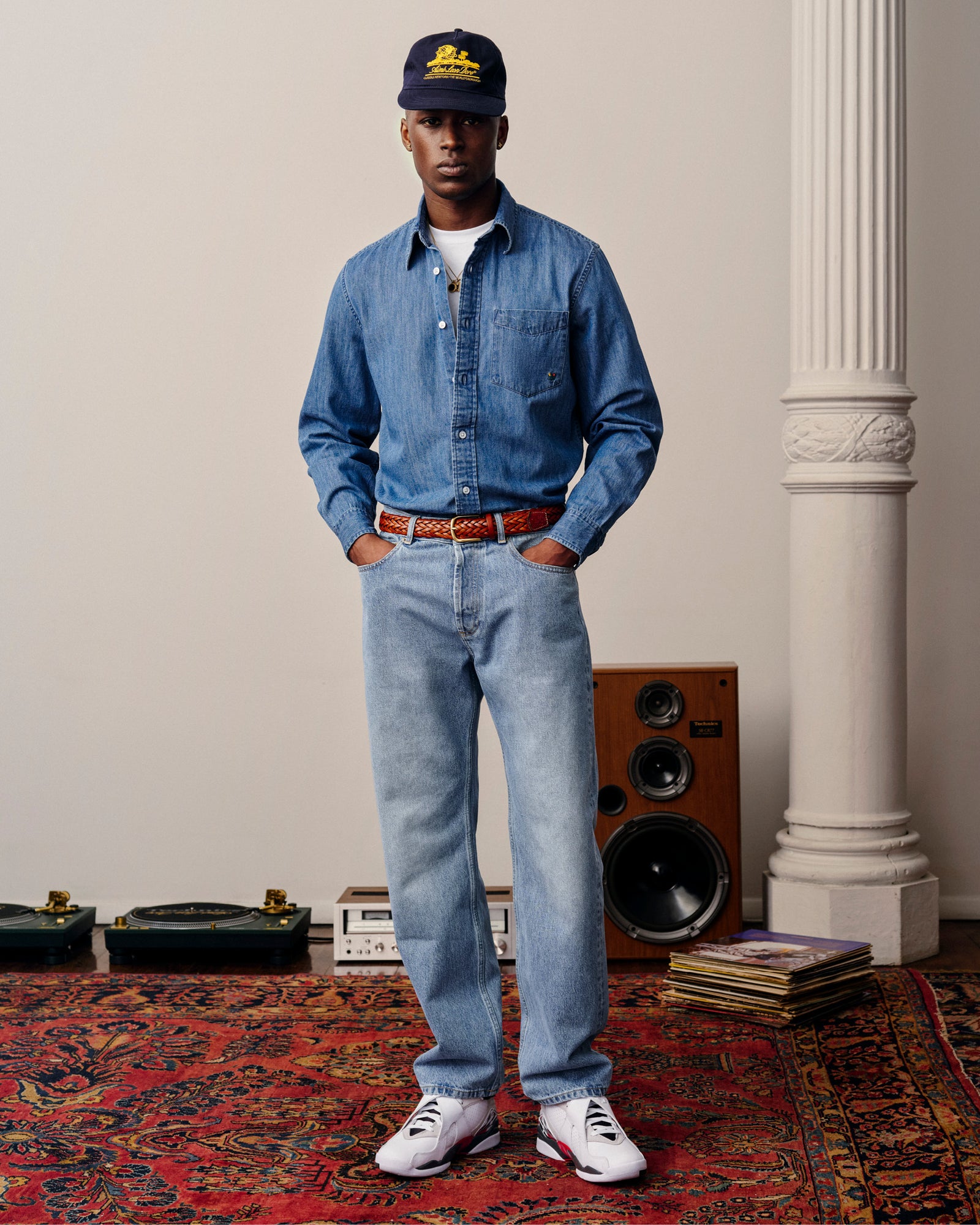 A man in ALD Straight Fit Denim stands on a red rug with speakers and records behind him.