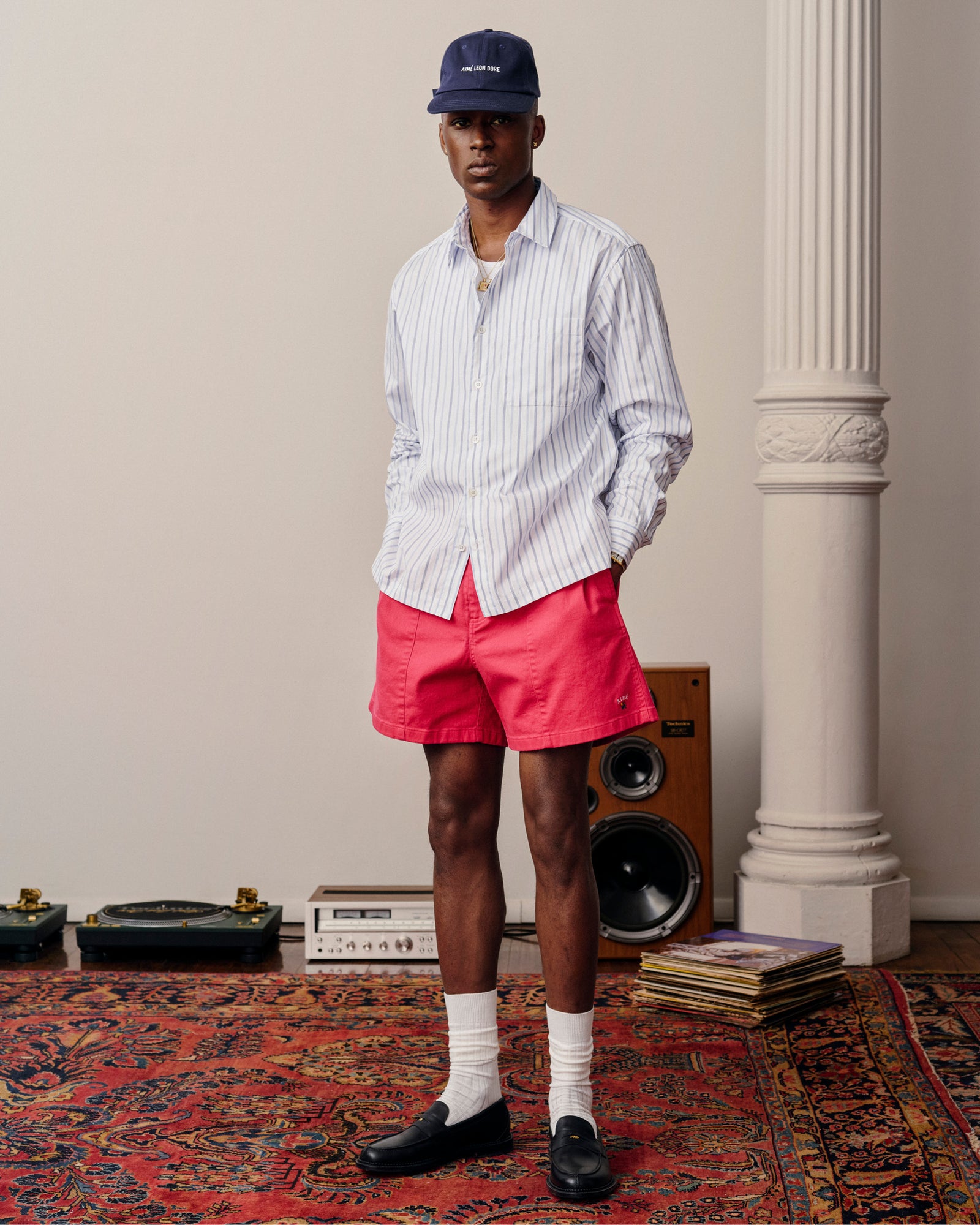 A man in a Poplin Casual Shirt, red shorts, and a cap stands on a patterned rug near speakers and vinyl records.