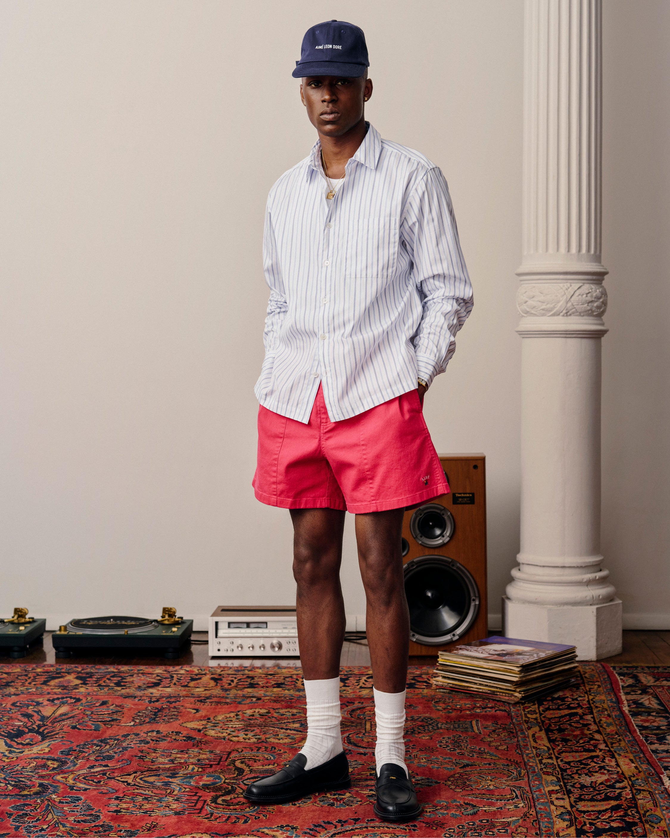 A man in a Poplin Casual Shirt, red shorts, and a cap stands on a patterned rug near speakers and vinyl records.