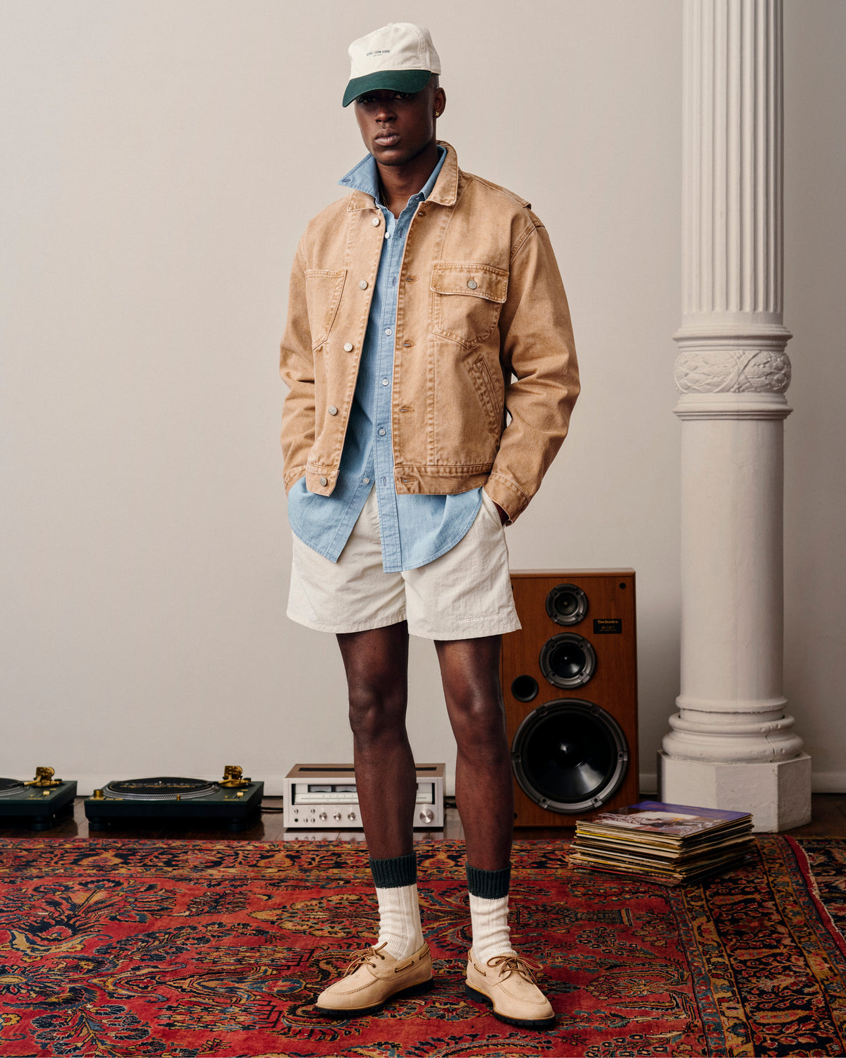 A man in a tan jacket, denim shirt, light shorts, and Micro Logo Hat stands on a patterned rug near speakers and records.