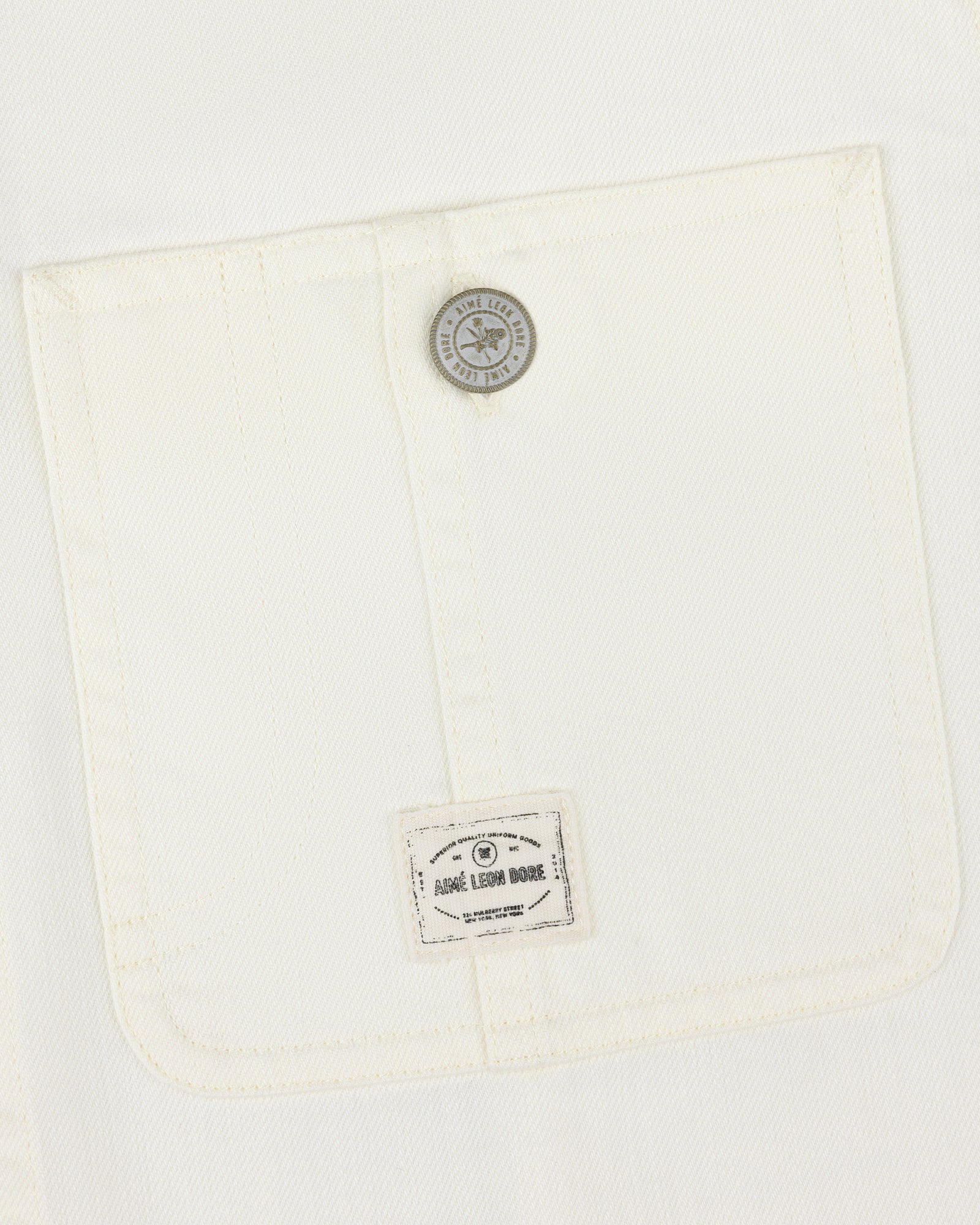 Close-up of a white Carpenter Overshirt pocket with a button and small Aimé Leon Dore label at the bottom.