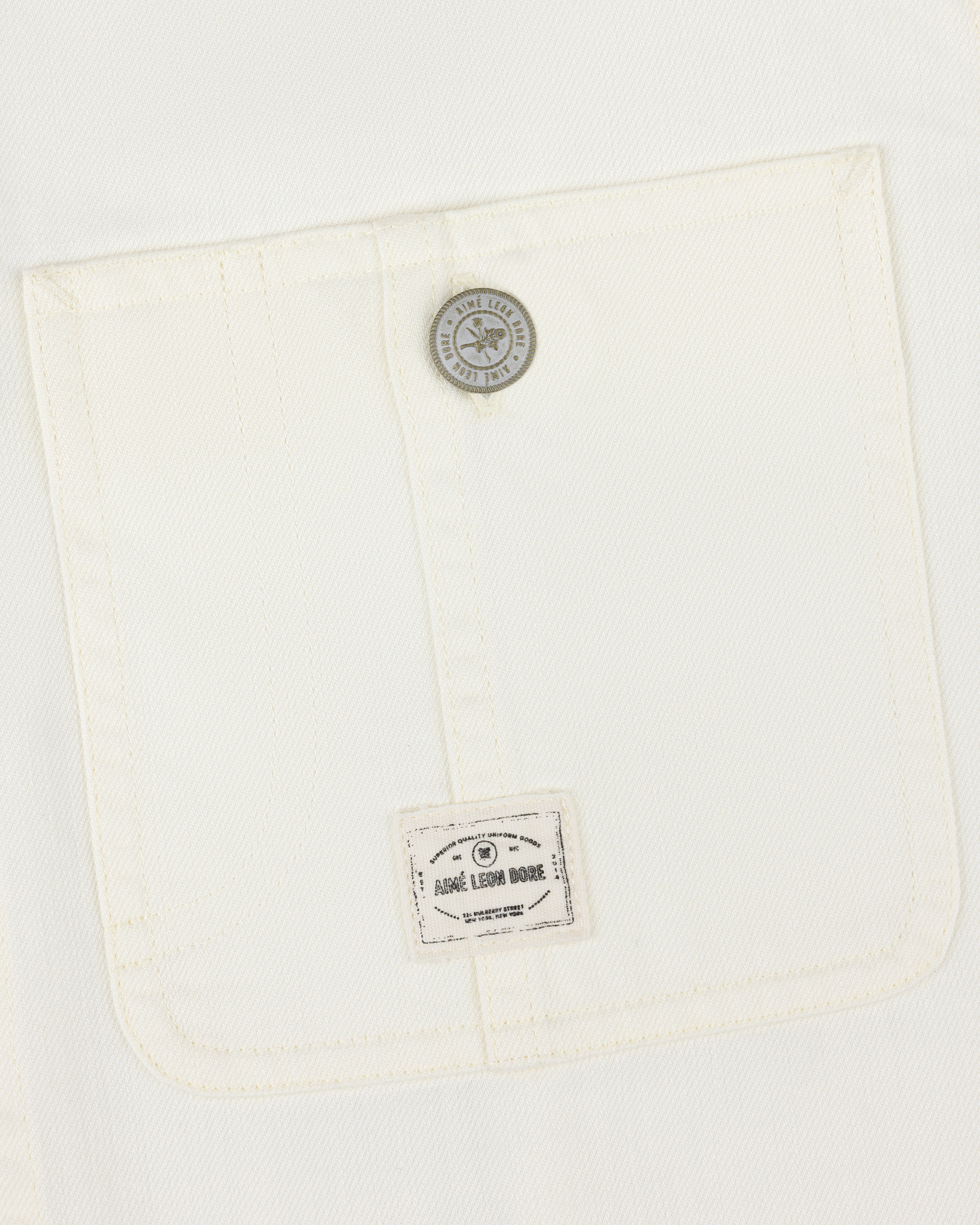 Close-up of a white Carpenter Overshirt pocket with a button and small Aimé Leon Dore label at the bottom.