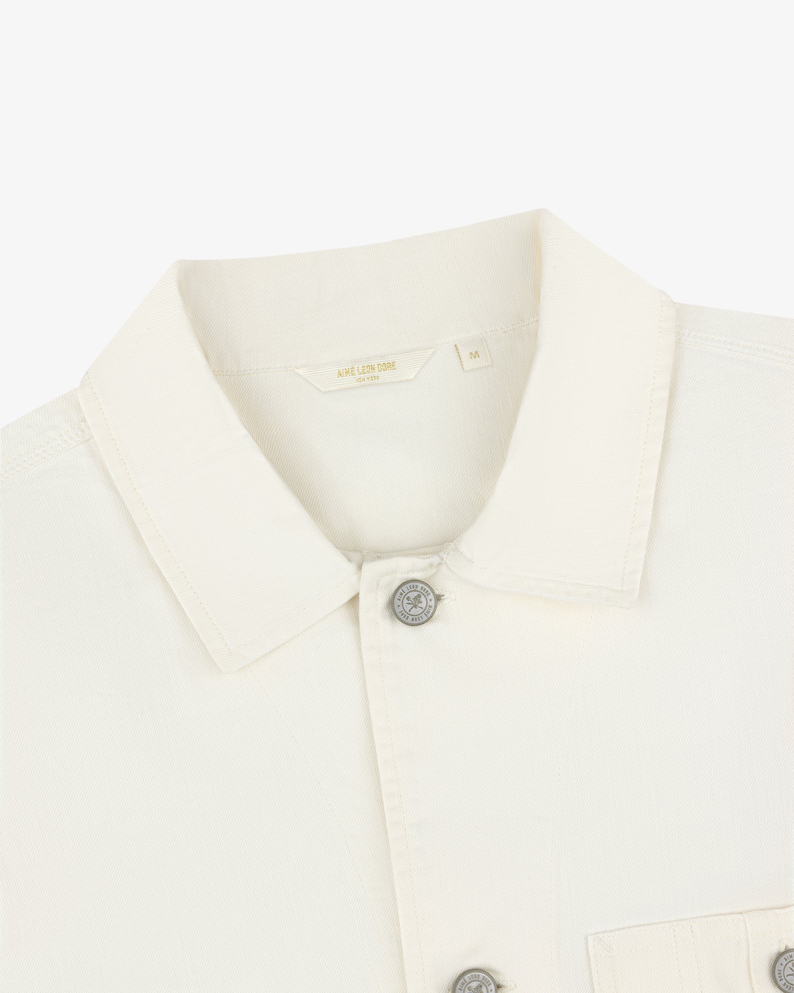 Close-up of a Carpenter Overshirt in off-white, with silver buttons, chest pocket, and size M label inside the collar.
