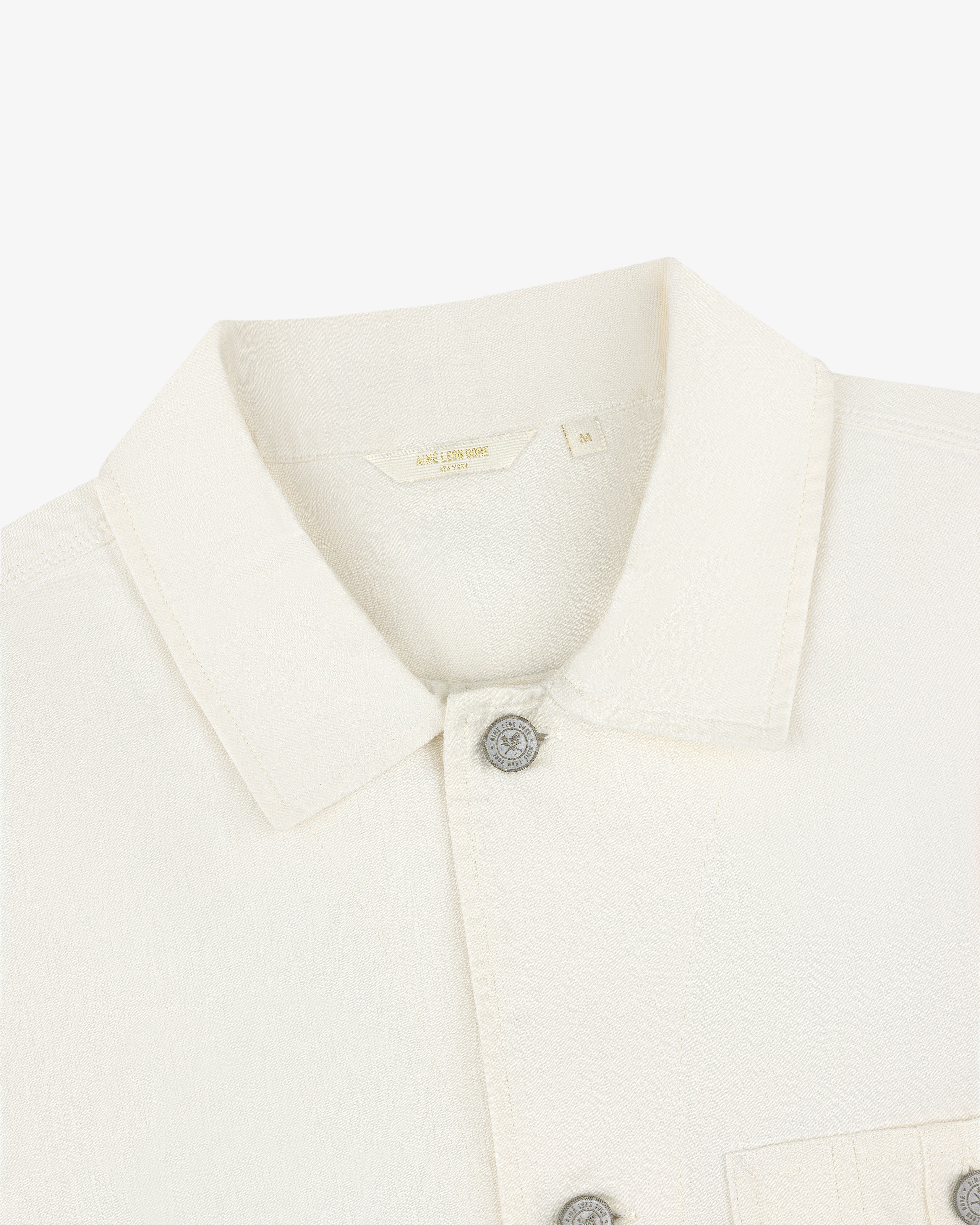 Close-up of a Carpenter Overshirt in off-white, with silver buttons, chest pocket, and size M label inside the collar.